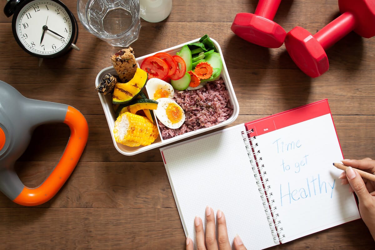 Person writing on a notebook with dumbbells and a lunch box on a table